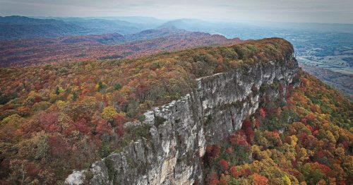 Aerial view of a rocky cliff surrounded by vibrant autumn foliage in a mountainous landscape.