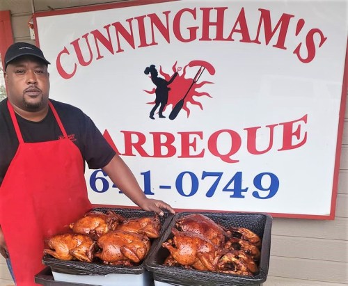A man in a red apron stands in front of a barbecue sign, holding two trays of seasoned cooked chickens.