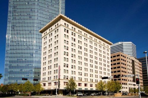 A historic white building stands next to modern skyscrapers under a clear blue sky. Trees line the street below.