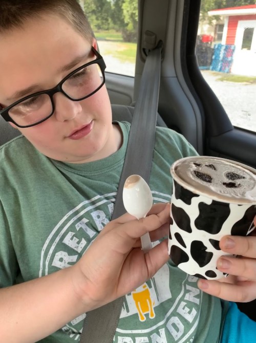 A boy in a car holds a spoon and a cup of ice cream with a cow print design, looking at the treat.