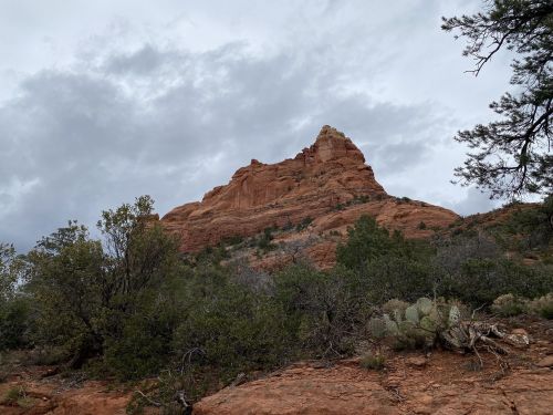 A rocky red mountain peak rises against a cloudy sky, surrounded by green shrubs and trees.