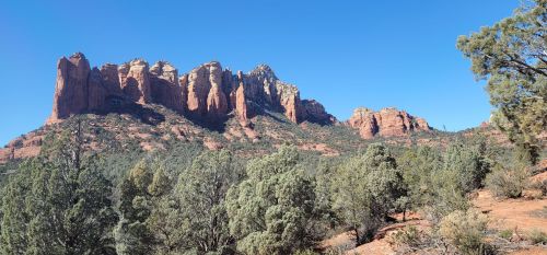 A panoramic view of red rock formations surrounded by green trees under a clear blue sky.