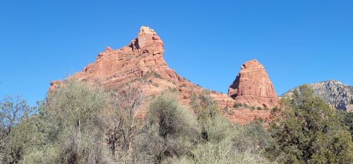 Red rock formations rise against a clear blue sky, surrounded by sparse vegetation in a desert landscape.