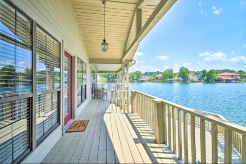 A sunny lakeside porch with wooden railing, overlooking calm water and lush greenery.