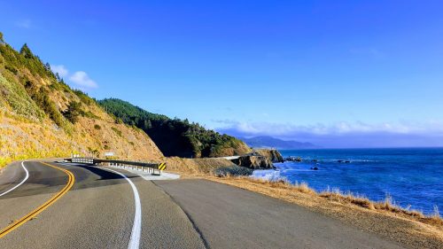 A winding coastal road curves along a scenic shoreline with blue ocean and green hills under a clear sky.