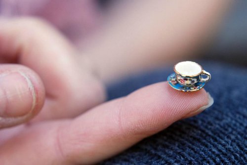 A person’s finger holds a tiny, decorative teacup and saucer, showcasing intricate details and vibrant colors.