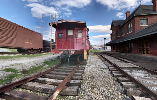 A red caboose on tracks beside a historic train station, with freight cars and a blue sky in the background.