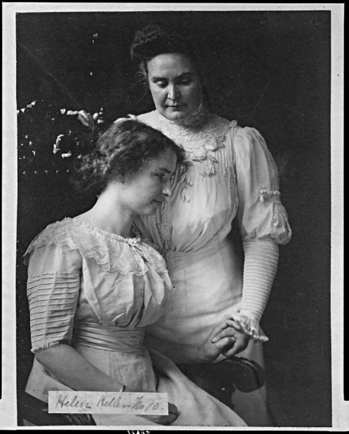 A black-and-white photo of two women, one seated and the other standing behind her, both in vintage dresses.