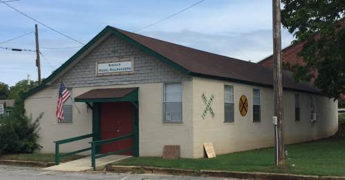 A single-story building with a green roof and door, featuring signs for a model railroad club and an American flag.