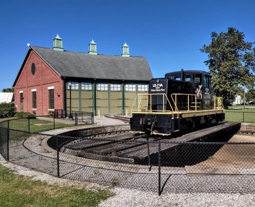 A black and yellow locomotive on a turntable in front of a green and red train depot under a clear blue sky.