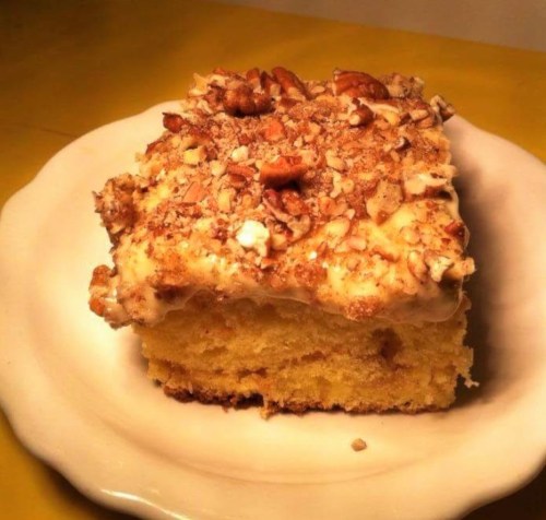 A slice of yellow cake topped with a creamy frosting and crushed nuts, served on a white plate.