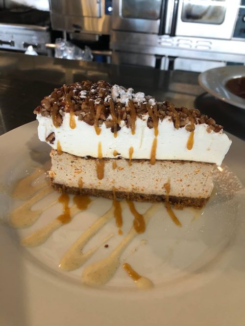 A slice of layered dessert with a graham cracker crust, creamy filling, whipped topping, and drizzled caramel.