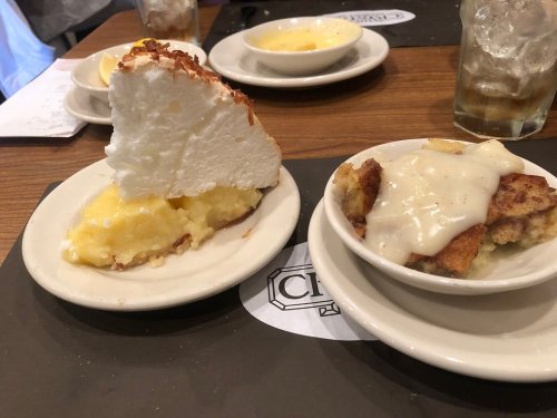 A slice of coconut cream pie next to a bowl of bread pudding topped with white sauce on a restaurant table.