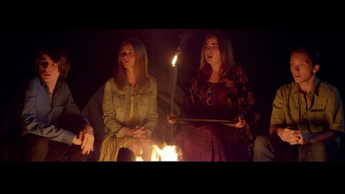 Four people sit around a campfire at night, listening intently, with a flickering flame illuminating their faces.