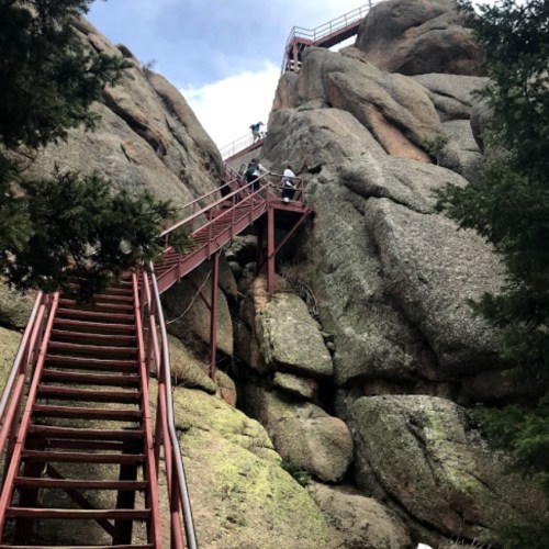 A steep metal staircase leads up a rocky cliff, surrounded by trees and a cloudy sky.