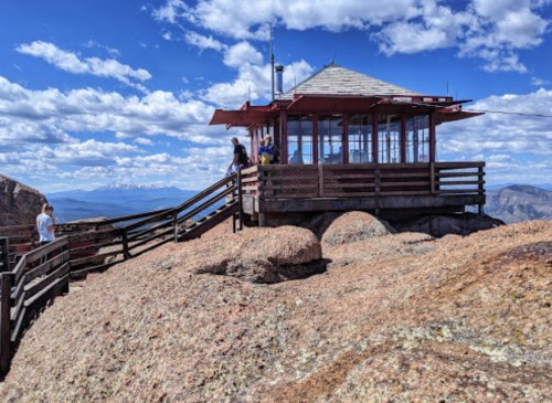 A lookout tower on a rocky peak, surrounded by clouds and mountains, with people enjoying the view.