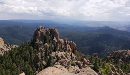 A panoramic view of rocky mountains and dense green forests under a cloudy sky.