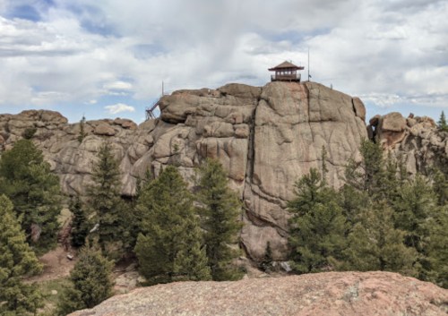 A rocky outcrop with a small lookout tower on top, surrounded by pine trees and a cloudy sky.