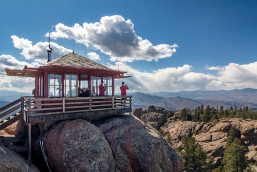 A lookout tower on a rocky outcrop, with people enjoying the panoramic mountain view under a blue sky with clouds.