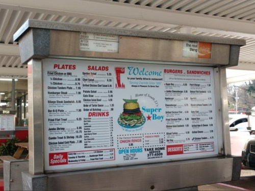 Menu board at a fast-food restaurant featuring burgers, sandwiches, salads, and drinks with prices listed.