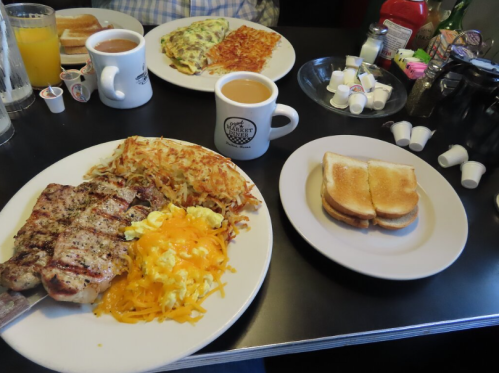 A breakfast spread featuring a plate of eggs, hash browns, grilled meat, toast, and coffee on a diner table.