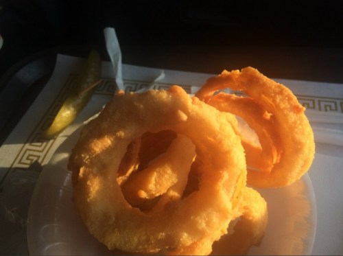 A plate of golden, crispy onion rings with a pickle slice in the background.