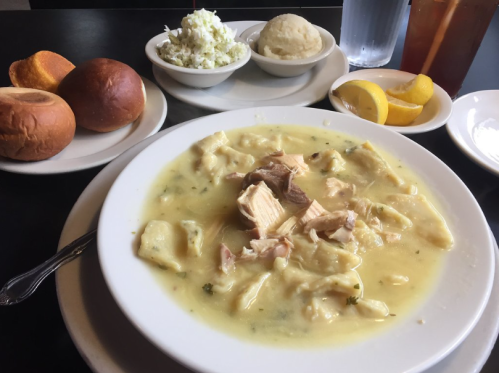 A bowl of chicken and dumplings with sides of coleslaw, mashed potatoes, rolls, and lemon wedges on a table.