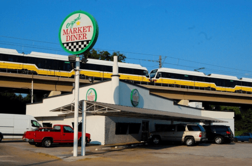 A diner with a green sign and a train passing overhead on an elevated track. Several cars are parked outside.