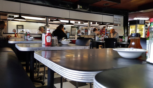 A diner interior with empty tables, ketchup bottles, and patrons seated in the background.