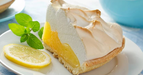 A slice of lemon meringue pie on a plate, garnished with a mint leaf and a lemon slice.