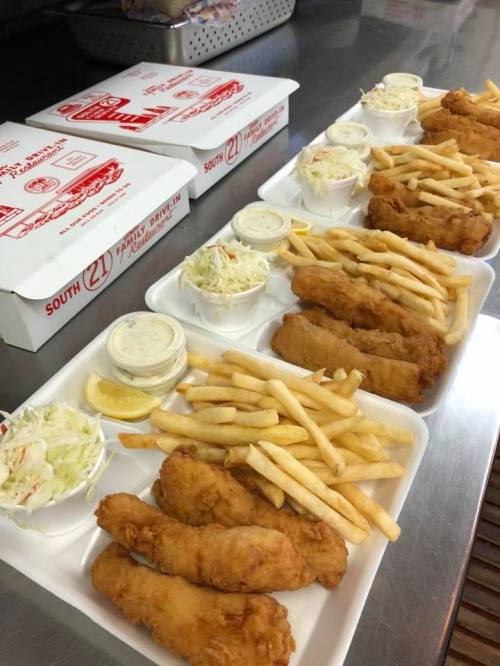 Two trays of fried fish, fries, coleslaw, and dipping sauces, with takeout boxes in the background.