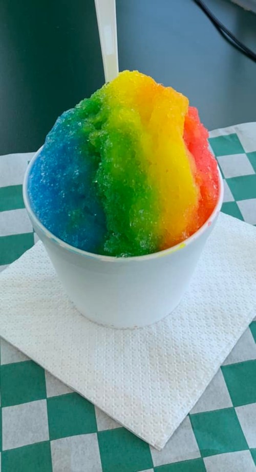 A colorful rainbow snow cone in a white cup, sitting on a green and white checkered napkin.