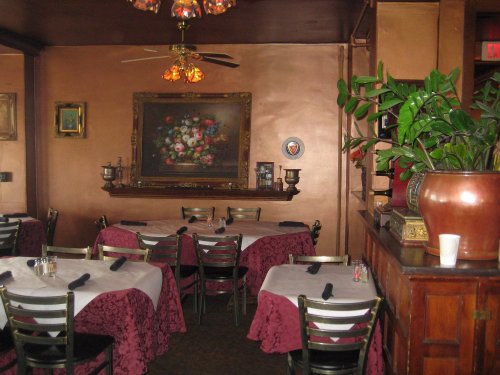Cozy restaurant interior with tables covered in white and burgundy linens, a floral painting, and a potted plant.