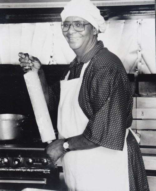 A smiling chef in a polka dot shirt and apron holds a rolling pin in a kitchen, with pots in the background.