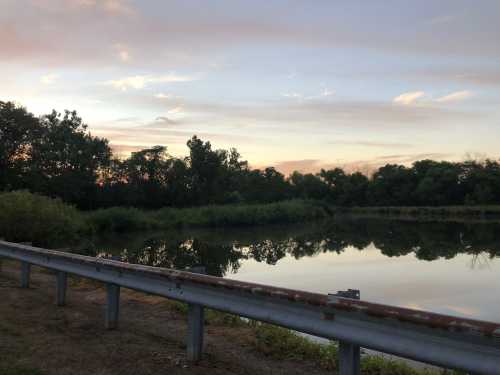 A serene landscape featuring a calm pond reflecting trees and a colorful sky at sunset, with a guardrail in the foreground.