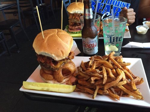 A towering burger with onion rings, fries, a pickle, and a beer on a table in a casual dining setting.