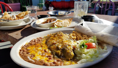 A plate of enchiladas topped with cheese, served with rice, beans, and a side salad, with additional dishes in the background.