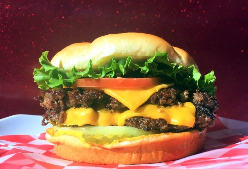 A close-up of a juicy cheeseburger with lettuce, tomato, and pickles on a soft bun against a sparkling red background.