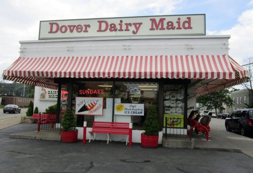 Exterior of Dover Dairy Maid, featuring a striped awning, red benches, and a sign advertising sundaes and soft serve ice cream.