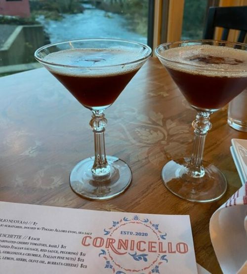 Two cocktails in elegant glasses on a wooden table, with a menu featuring "Cornicello" in the background.