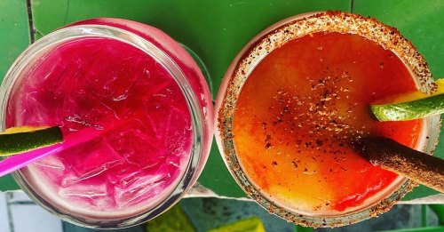 Two colorful cocktails on a green surface: one pink with ice and a straw, the other orange with a rim and lime garnish.