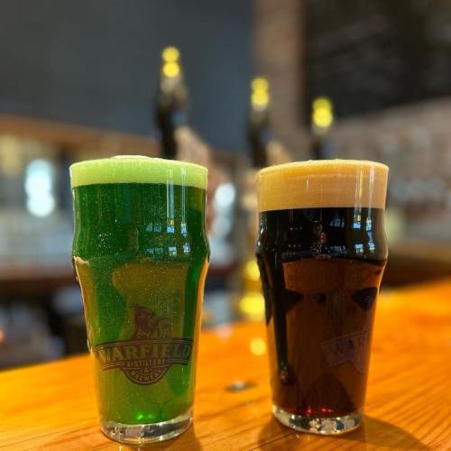 Two pint glasses on a wooden bar: one filled with green beer and the other with dark stout, both branded with a logo.