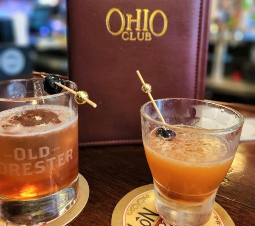 Two cocktails on a bar top, one in a glass with ice and a cherry, the other in a short glass, with a menu in the background.