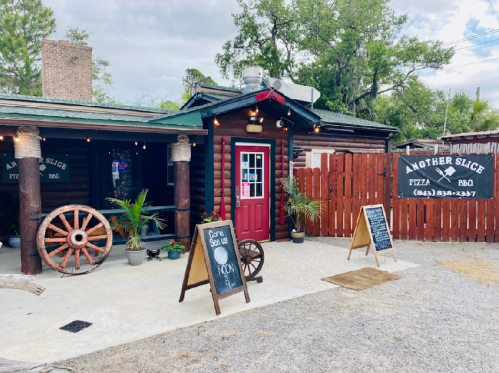 Exterior of "Another Slice" restaurant featuring a wooden facade, a red door, and signs for pizza and BBQ.