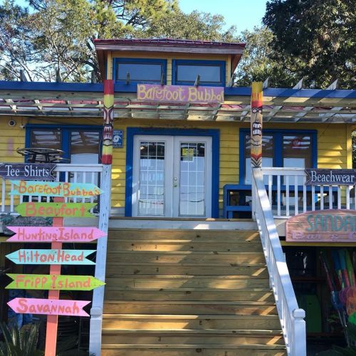 Colorful beach shop with a sign for "Barefoot Bubba," featuring stairs and directional signs to nearby locations.