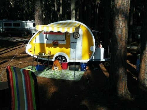 A bright yellow and white camper trailer set up in a wooded campsite, with a green mat and camping gear around it.