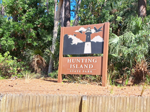 Sign for Hunting Island State Park featuring a lighthouse and birds, surrounded by trees and greenery.