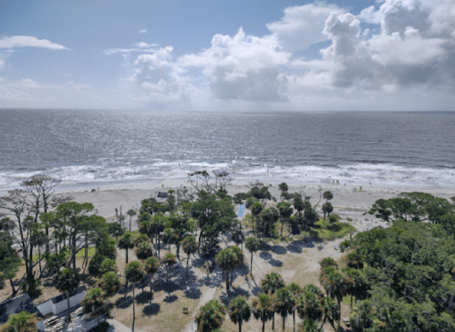 Aerial view of a beach with palm trees, gentle waves, and a cloudy sky in the background.