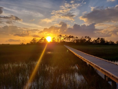 A wooden boardwalk stretches into a sunset over a tranquil marsh, with vibrant clouds and reflections in the water.