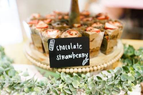 Chocolate strawberry desserts in clear cups, topped with strawberries, displayed on a wooden stand with greenery.